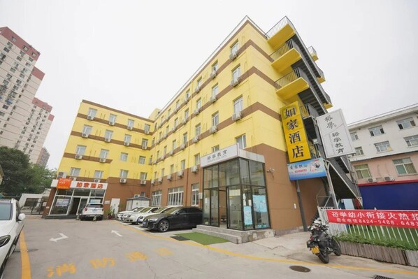 Un hôtel à plusieurs étages, avec une façade jaune, une entrée bien visible et un parking où sont garées des voitures.