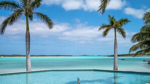 En pool med krystalklart blåt vand, omgivet af palmer, med udsigt til havet og landskabet i det fjerne.