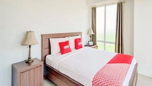 Habitación de hotel con cama de madera, ropa de cama blanca, almohadas rojas, una lámpara de noche y vistas al exterior a través de una ventana con cortinas.