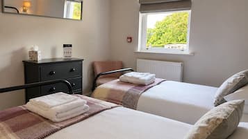A bedroom with two beds, a bedside table, a dresser, a window with a view of greenery, and a wall-mounted mirror.