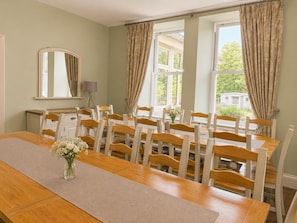 Un espace repas avec une longue table en bois et des chaises blanches, un miroir au mur et une vue sur la verdure à l’extérieur.