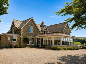 A stone house with a large glass conservatory, surrounded by gravel and greenery.