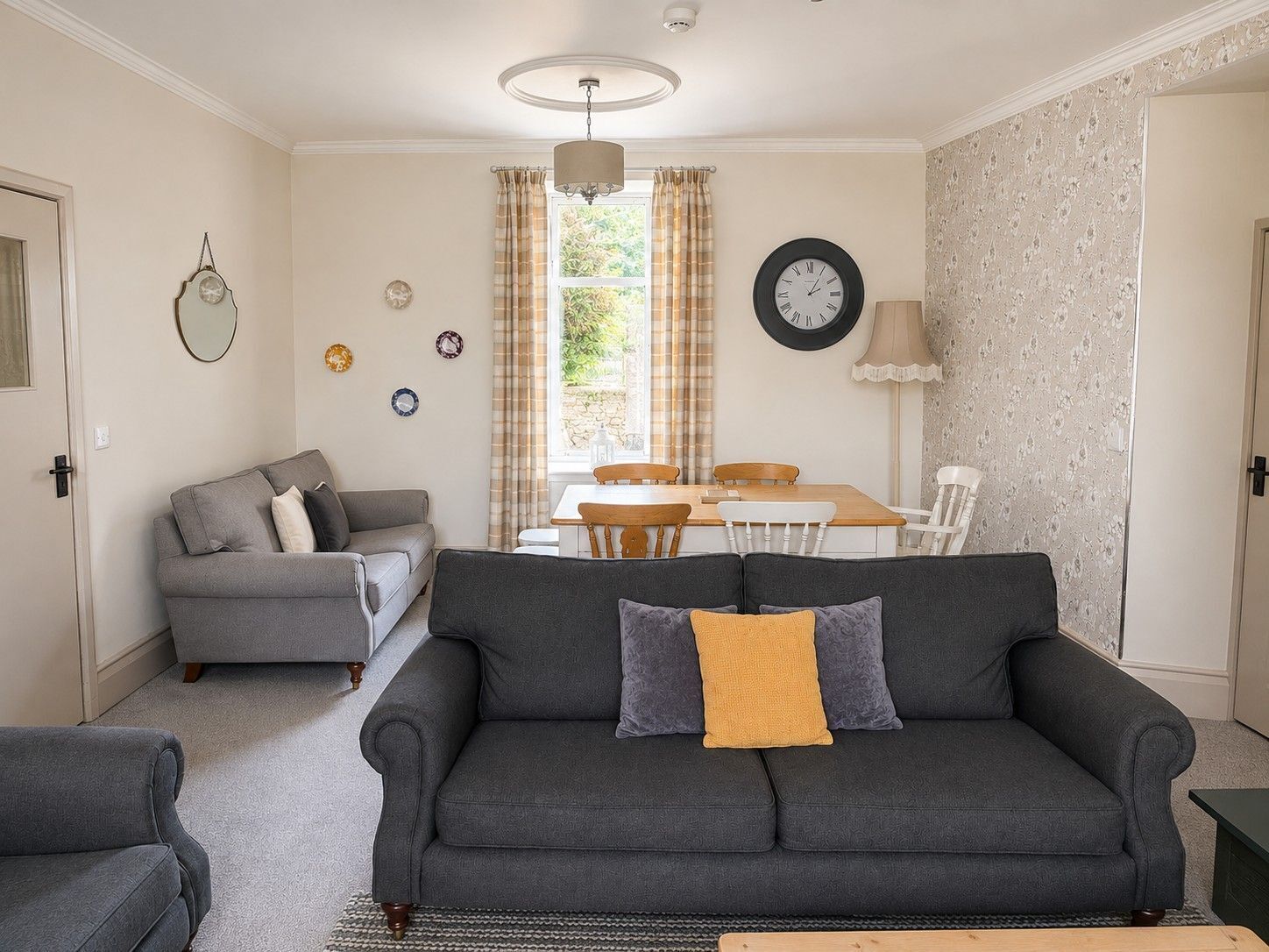 Ein Wohnzimmer mit grauem Sofa, einem kleinen grauen Sessel, einem Holztisch und einem Esstisch mit Stühlen. An der Wand hängt eine Uhr.