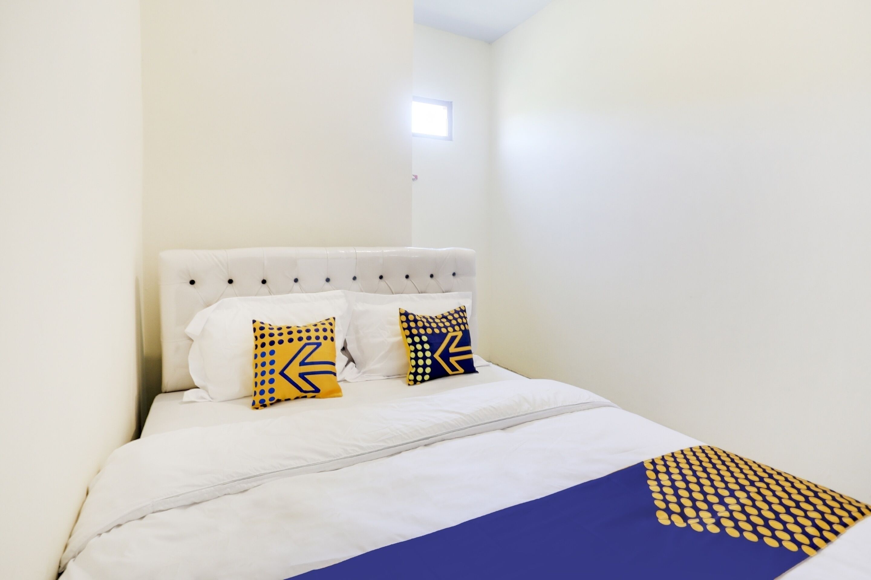 Una cama bien hecha con ropa blanca y dos almohadas decorativas, una pequeña ventana encima de la cama y una pared blanca lisa.