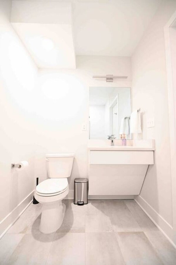 Une petite salle de bain blanche, avec des toilettes, une poubelle et un lavabo surmonté d’un miroir.