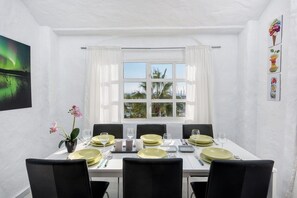 Un comedor con una mesa lista para cuatro personas, paredes blancas, un ventanal con cortinas y vistas a palmeras.