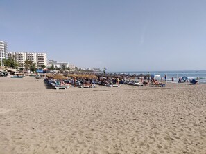 Una playa con tumbonas y sombrillas, cielo despejado y edificios al fondo.