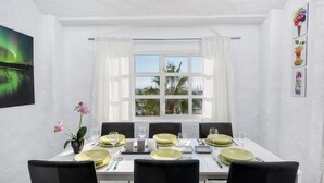 Un coin repas avec une table blanche dressée pour six personnes, des chaises noires et une vue sur les palmiers à travers une fenêtre ornée de rideaux blancs.
