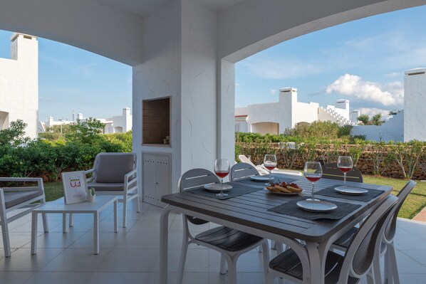 Un espace extérieur aménagé pour prendre les repas, avec une table dressée pour deux personnes, entourée de chaises blanches, d’une petite table et d’un barbecue.