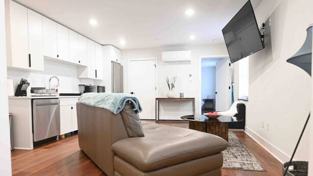 A modern living room with a sofa, a flat-screen TV mounted on the wall, and a kitchen area in the background. - Unit 102 · Near Yale and Downtown Newly Renovated (New Haven)