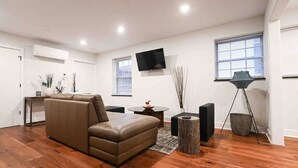A modern living room with a brown leather sofa, a flat-screen TV mounted on the wall, a round coffee table, and a floor lamp. - Unit 102 · Near Yale and Downtown Newly Renovated (New Haven)