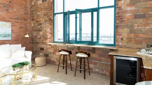 A spacious room with a brick wall, a large window, a white sofa, two bar stools, a small round table with a plant, and a cabinet with a television.