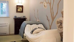 A room with a tatami mat floor, a low bed with a white headboard, a wall with a floral mural, a radiator, a framed picture on the wall, and a small table with a lamp. - Wabi Sabi B&B - a Japanese Sleep Oasis in Belgium (Peer)