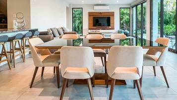 Een moderne eetruimte met een grote tafel en stoelen, een aan de muur bevestigde televisie en grote ramen met uitzicht op groen.
