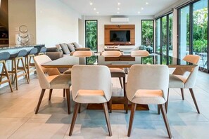 Un espace de repas moderne, doté d’une grande table et de chaises, d’un téléviseur fixé au mur et de grandes fenêtres offrant une vue sur la verdure.