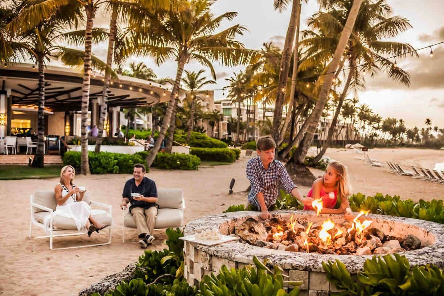 A group of people around a fire pit in a resort setting with palm trees and outdoor seating.