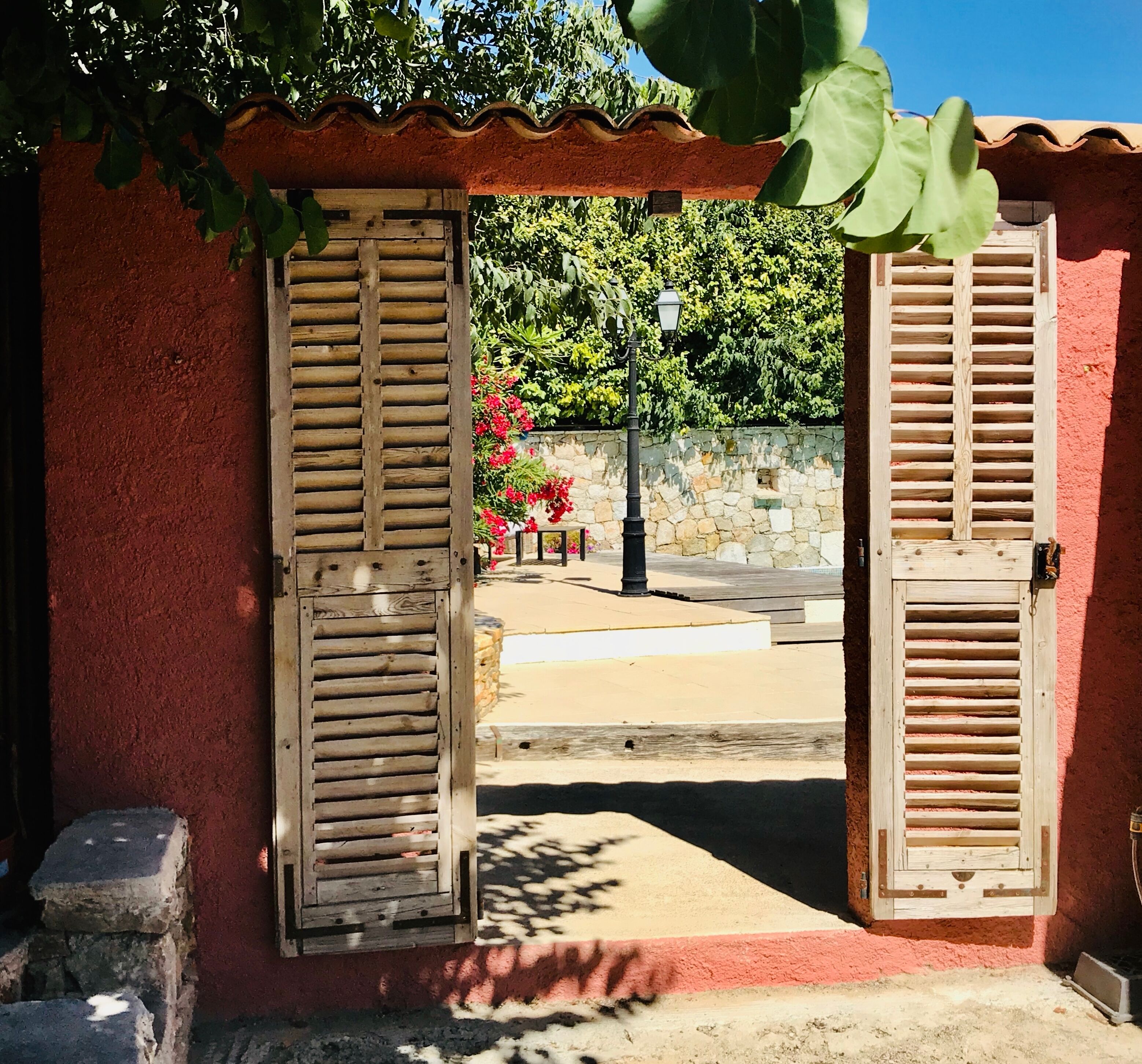 Une porte rustique avec des volets en bois, un mur de pierre et une vue sur la rue, avec un lampadaire et des fleurs.