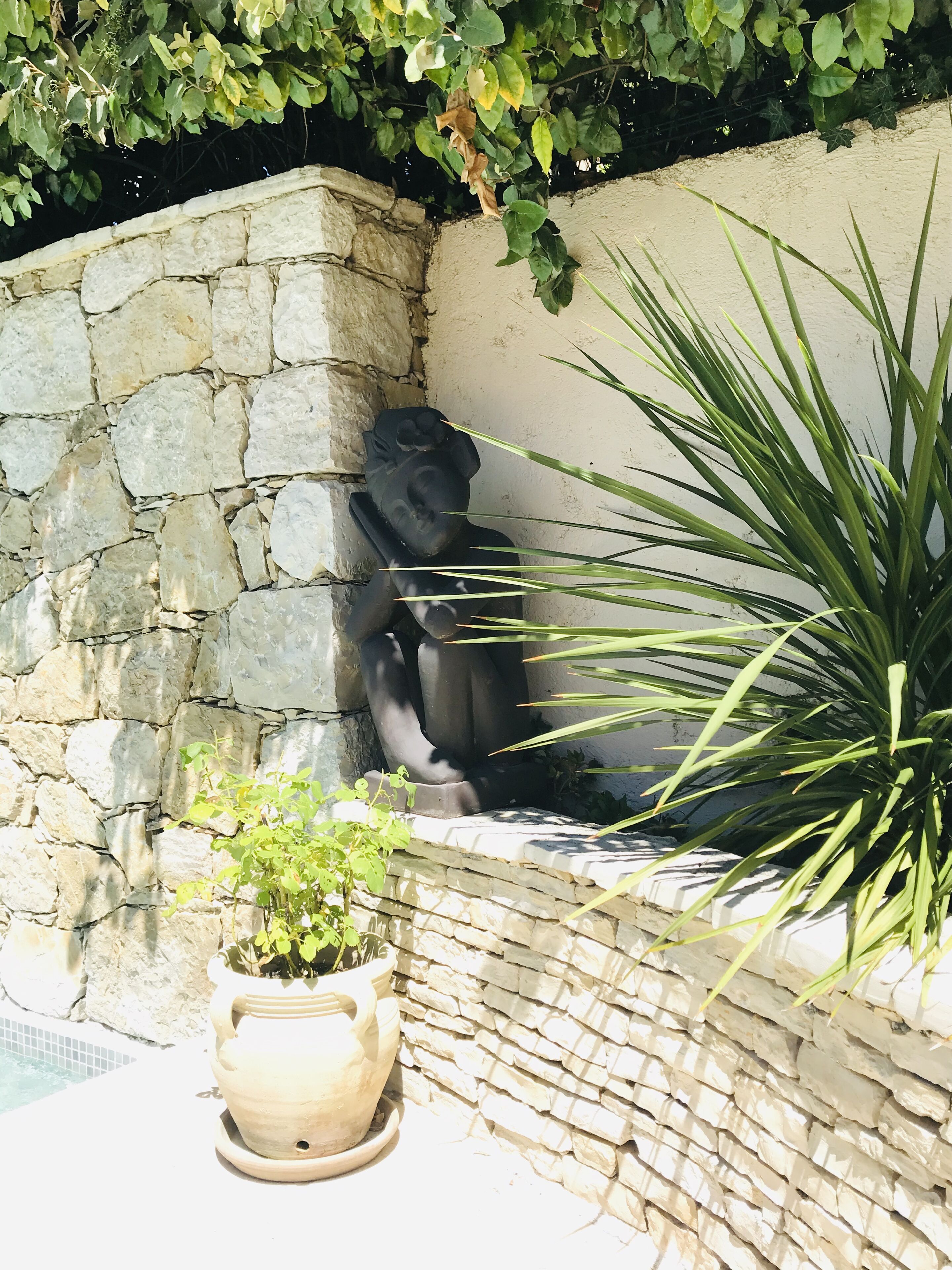 Un muro de piedra con una estatua, una planta en maceta y una zona junto a la piscina.