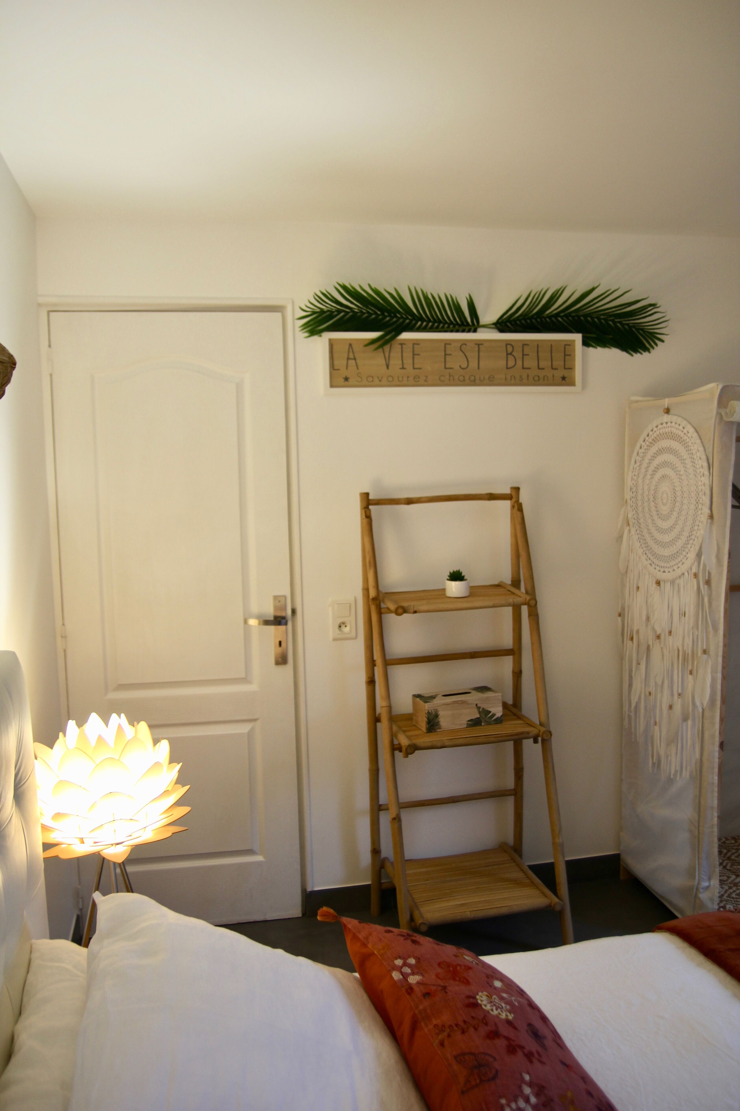 A bedroom with a bed, a bedside lamp, a wooden ladder shelf, a dreamcatcher, and a sign above the door.