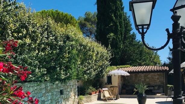 A swimming pool with a stone wall, wooden deck, and a small building with a tiled roof. - Villa Mathis (Mouans-Sartoux)