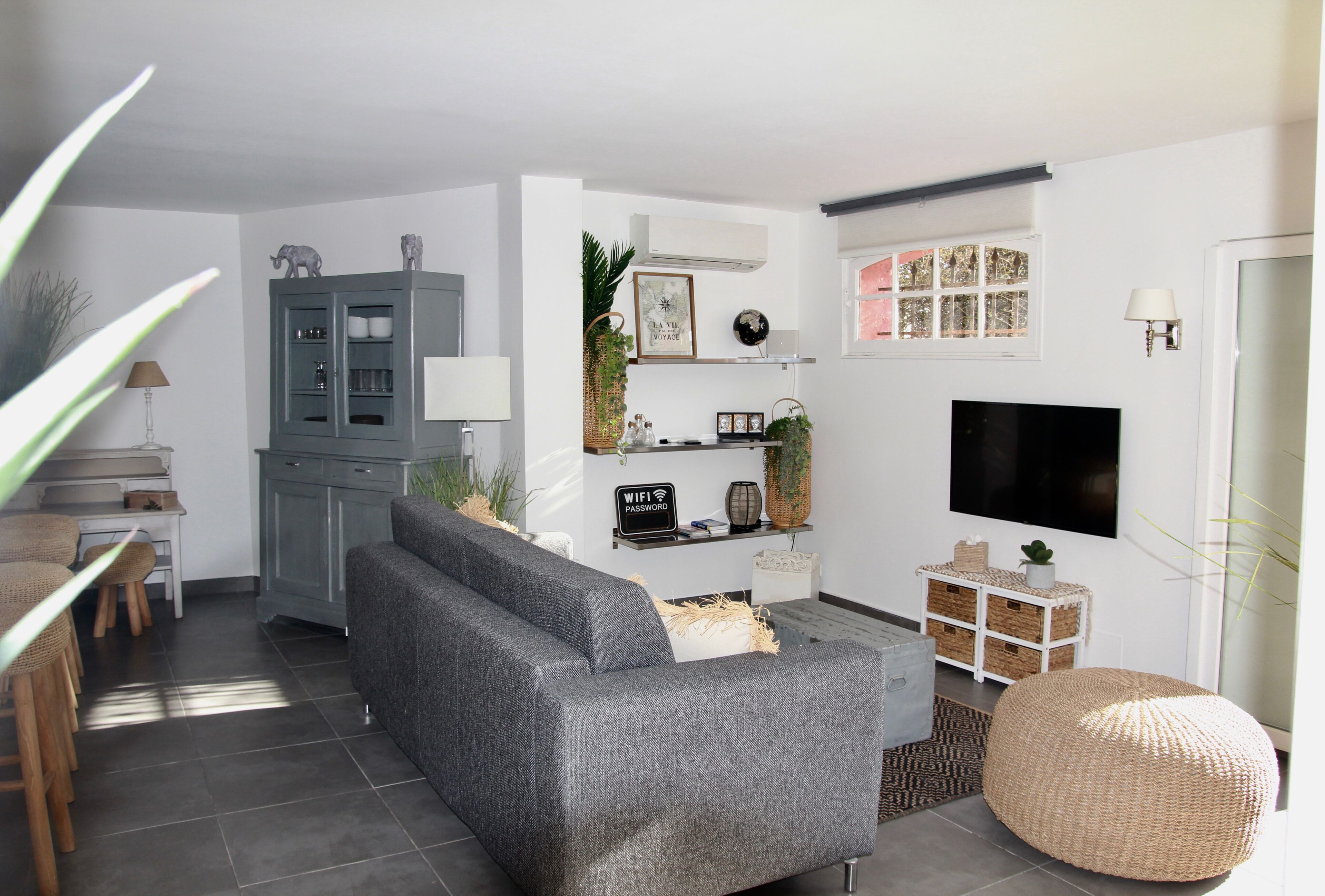 Un salon moderne avec un canapé gris, un pouf rond en fibres tressées, un téléviseur à écran plat et un meuble gris.