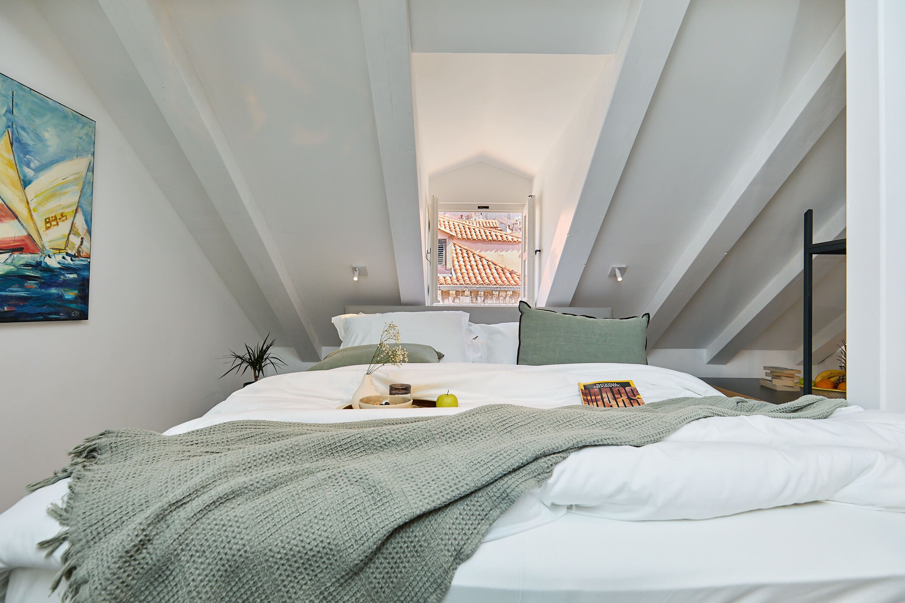 Une chambre à coucher moderne avec un plafond en pente, un lit recouvert de draps blancs, une couverture verte en tricot, une petite plante et un tableau représentant un voilier.