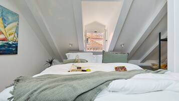 Une chambre à coucher moderne avec un plafond en pente, un lit recouvert de draps blancs, une couverture verte en tricot, une petite plante et un tableau représentant un voilier.
