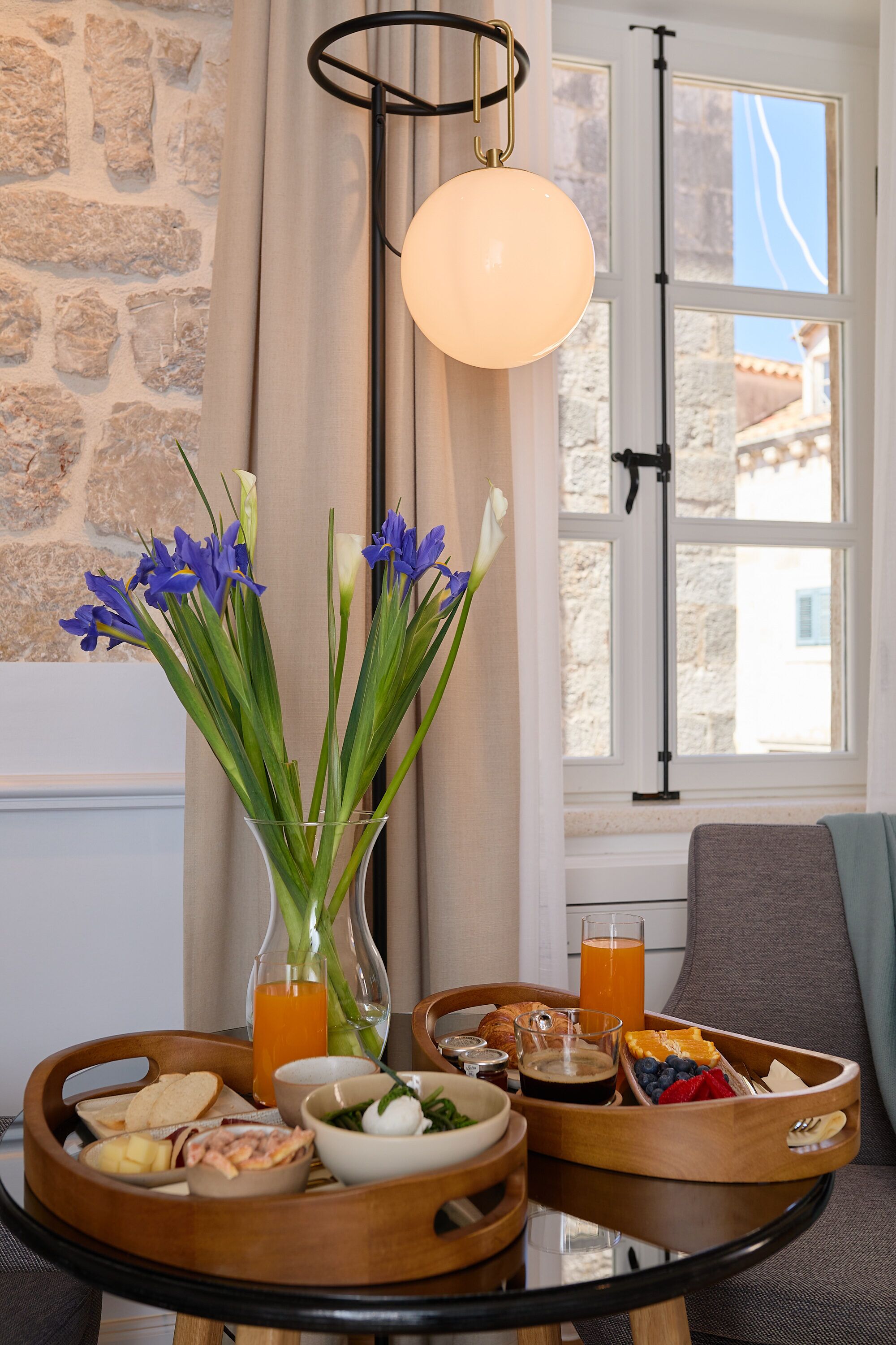 A breakfast table with a variety of food items, a vase with flowers, and a view of a stone wall and window.