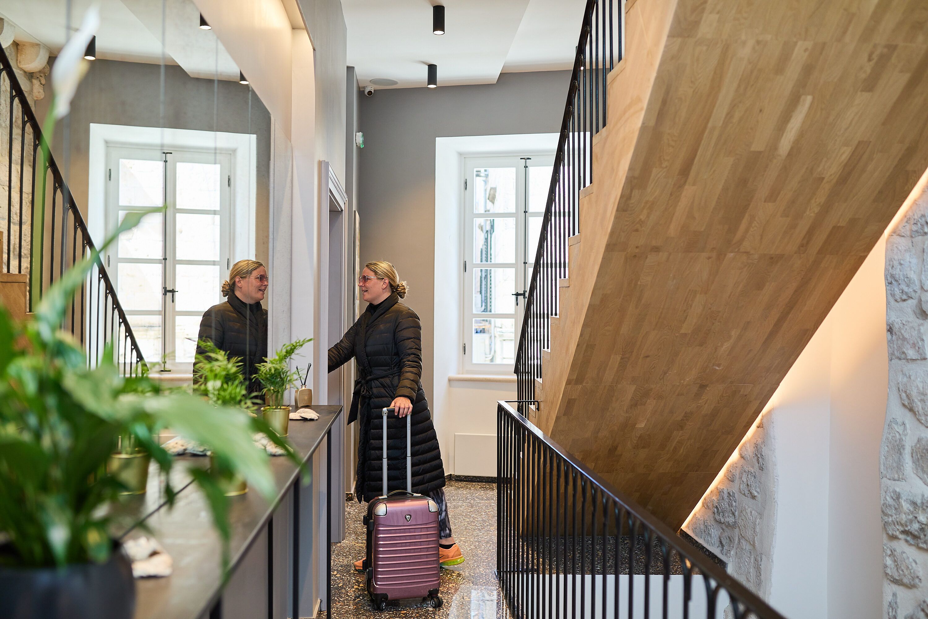 A hotel lobby with a staircase, potted plants, and a person with a suitcase.