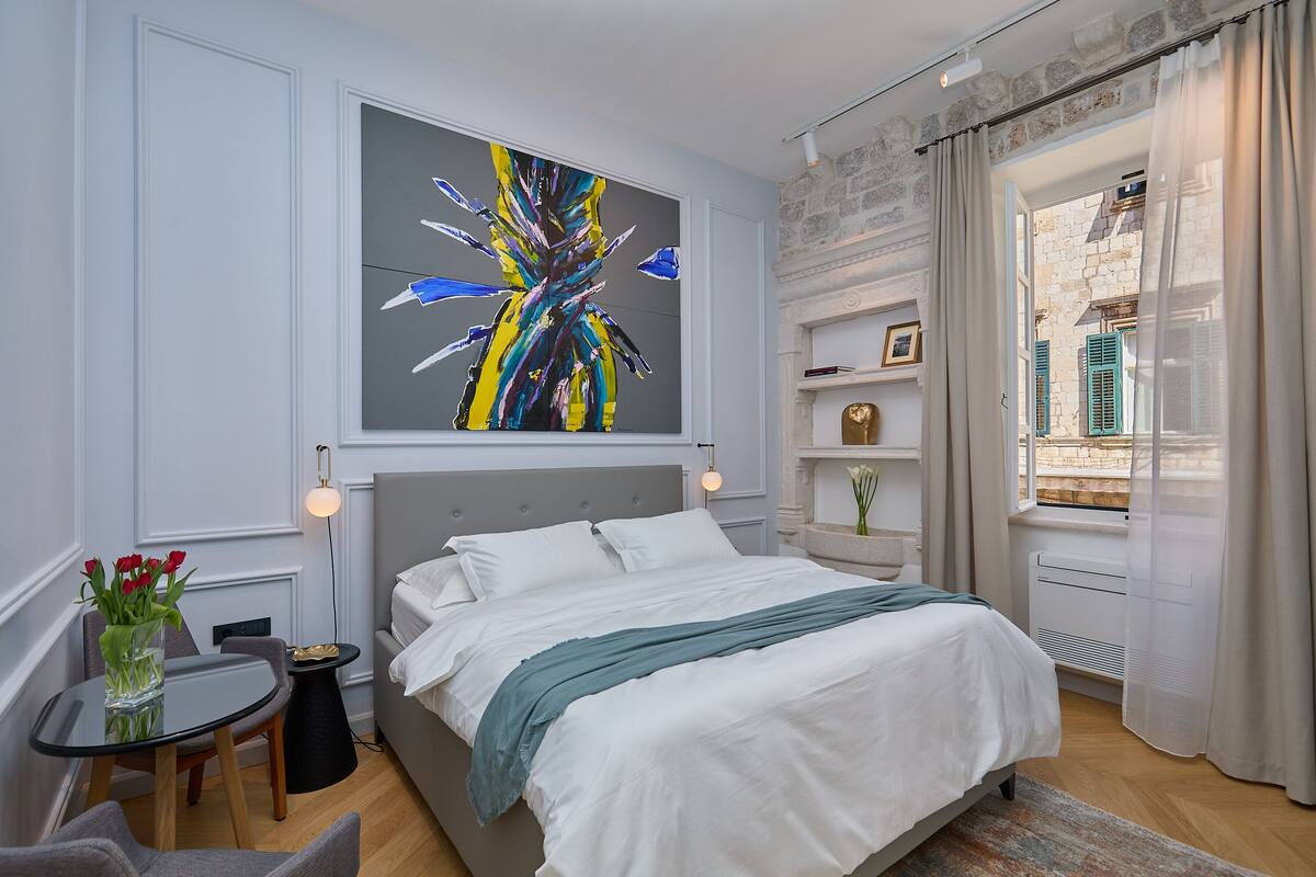 A modern bedroom with a large bed, a small table with a vase of flowers, a wall-mounted painting, and a view of the outside through a window with curtains.