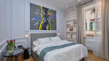 A modern bedroom with a large bed, a small table with a vase of flowers, a wall-mounted painting, and a view of the outside through a window with curtains.