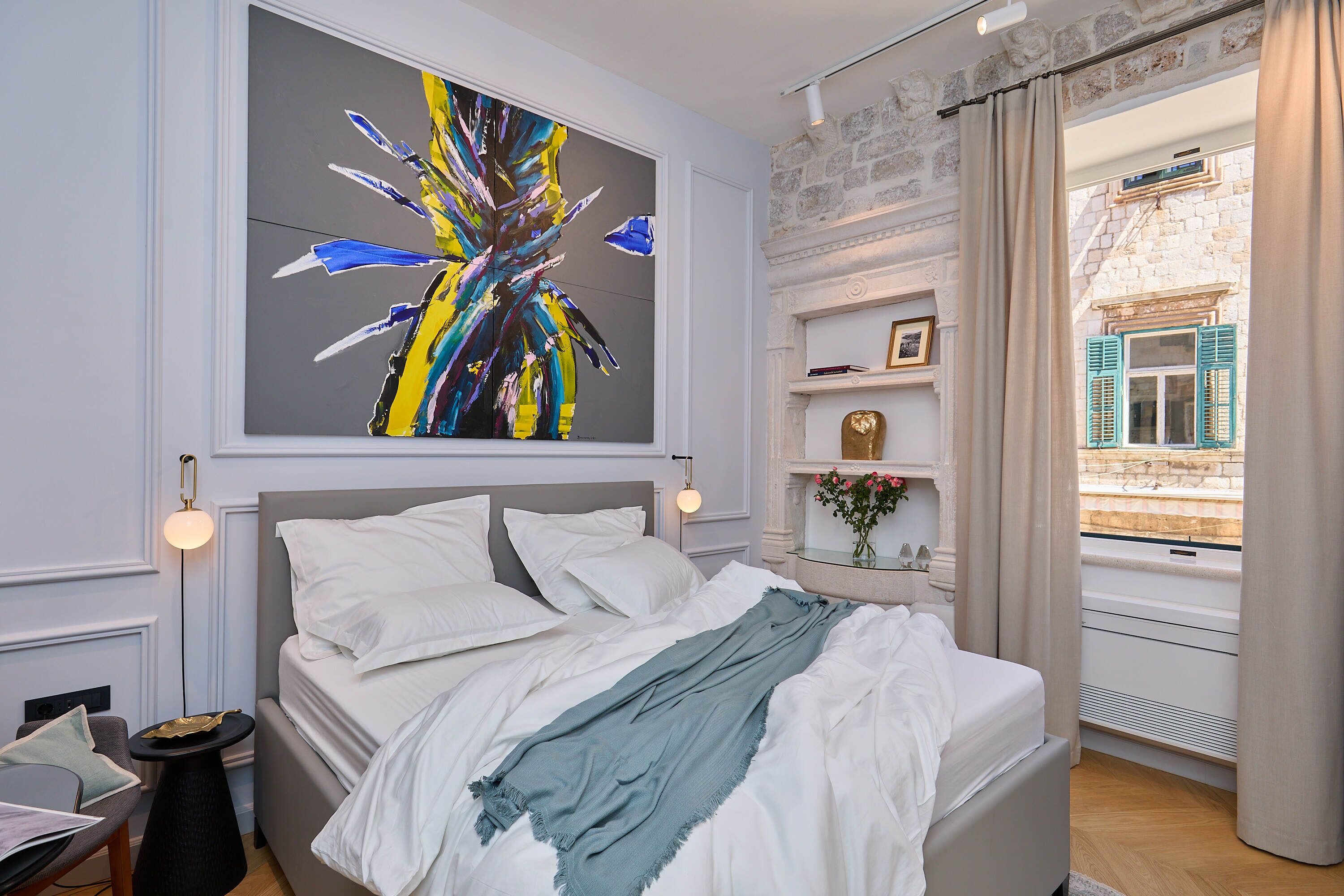 Quarto moderno com uma cama grande, uma mesa de cabeceira, um quadro e vista para um edifício pela janela.