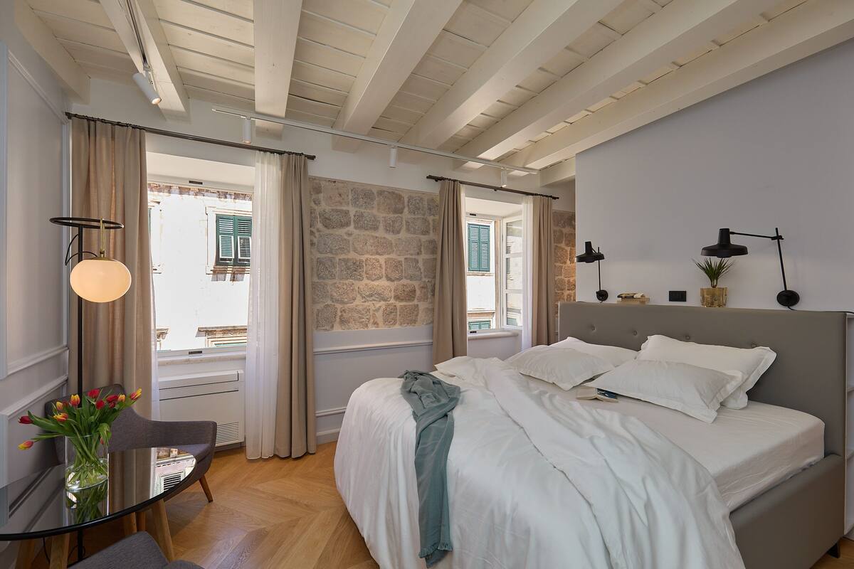 A bedroom with a bed, a chair, a table, a vase with flowers, and a stone wall.