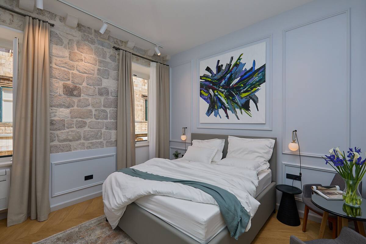 A modern bedroom with a stone wall, a large bed, a painting, and a view of the outside.