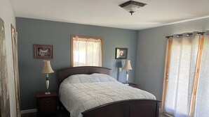 A bedroom with a wooden bed, two bedside tables with lamps, a window with sheer curtains, and a ceiling light fixture.