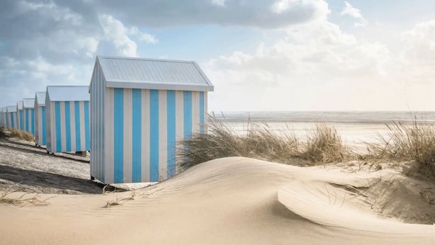 Een rij strandhutten met blauw-witte strepen, gelegen aan een zandstrand met duinen op de voorgrond.