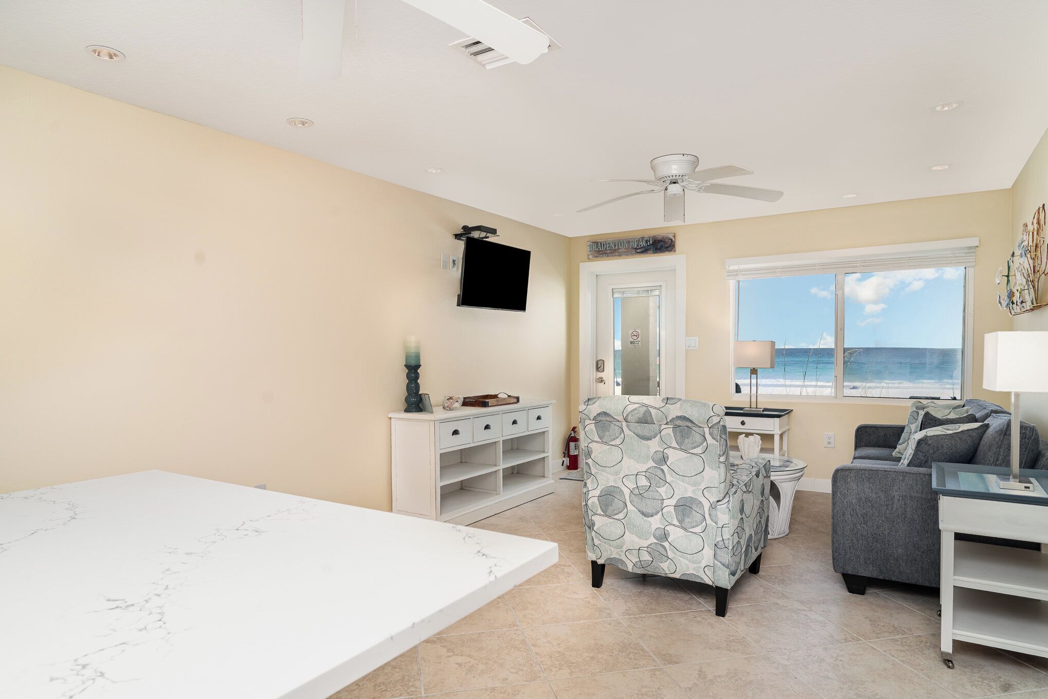 Un soggiorno spazioso con una grande finestra vista mare, una ventola a soffitto, una TV a schermo piatto montata a parete e una zona salotto accogliente con una poltrona fantasia e un divano grigio.