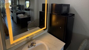A bathroom with a large mirror, a sink, a black refrigerator, and a chair. - I-26 Inn (Johnson City)