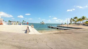 A beachside view with a concrete walkway leading to a dock, palm trees, and a clear blue sky. - Seas The Day in Cudjoe Key (Cudjoe Key)