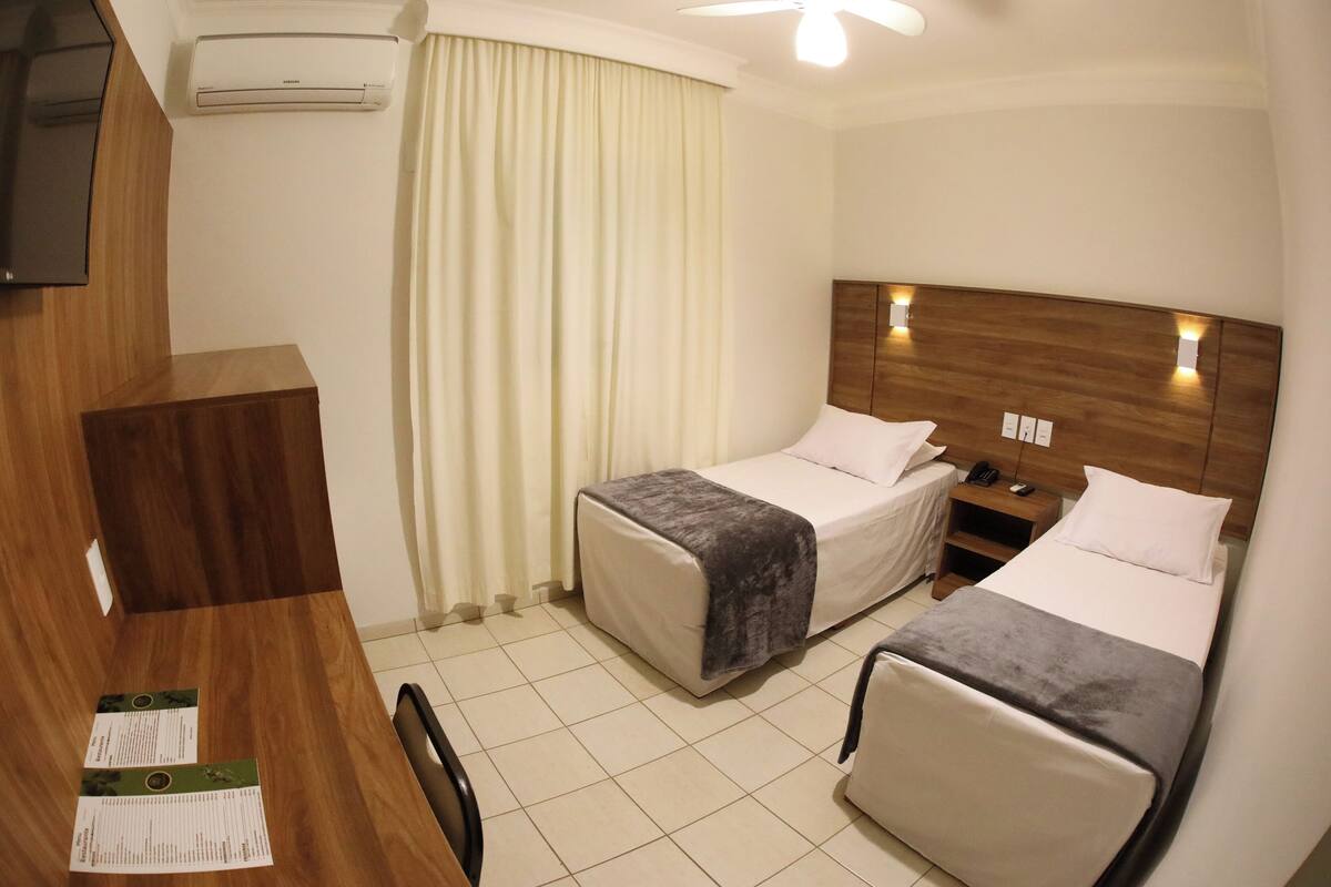Ett hotellrum med två sängar, ett skrivbord, en stol, en TV, en luftkonditionering och en gardin.
