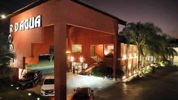 Un edificio con un cartel que dice "R D'AQUA" de noche, con un diseño moderno, fachada roja, una escalera visible y un estacionamiento con coches.