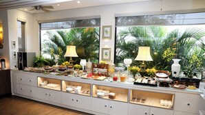 Una mesa de buffet con diversos alimentos y bebidas, un amplio ventanal con vista a palmeras y un ventilador de techo.