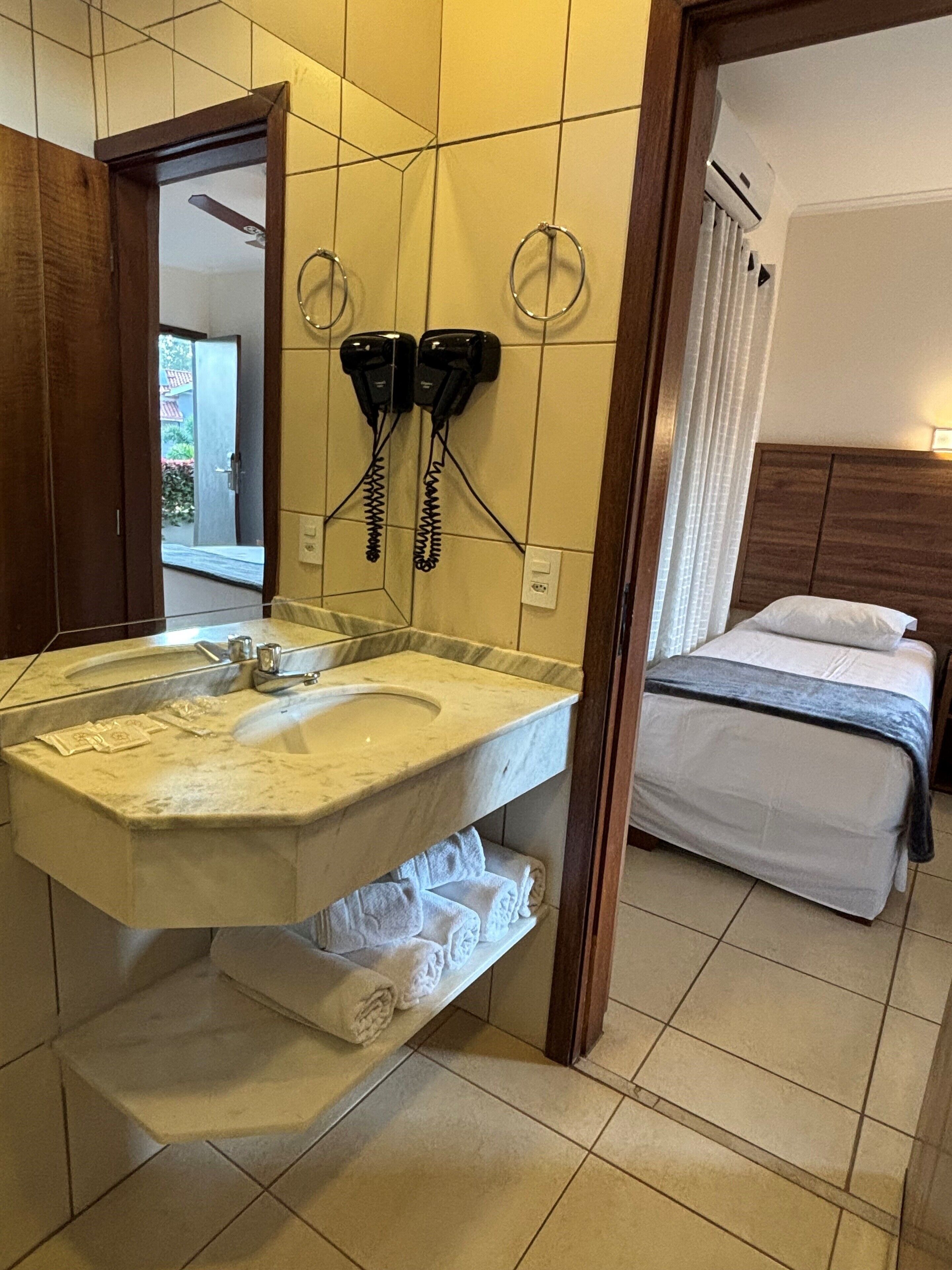 A bathroom with a marble sink, two hair dryers mounted on the wall, a bed with a wooden headboard, and a view of the outside through an open door.