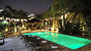 Zona de piscina nocturna con sillones de descanso y una estructura de techo de paja.