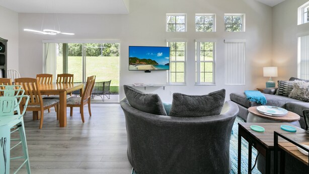 A spacious living area with a grey sofa, a dining table with chairs, and a television mounted on the wall.