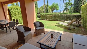 Un patio con mobiliario de mimbre, una mesa y vistas a un jardín cuidado y al mar.