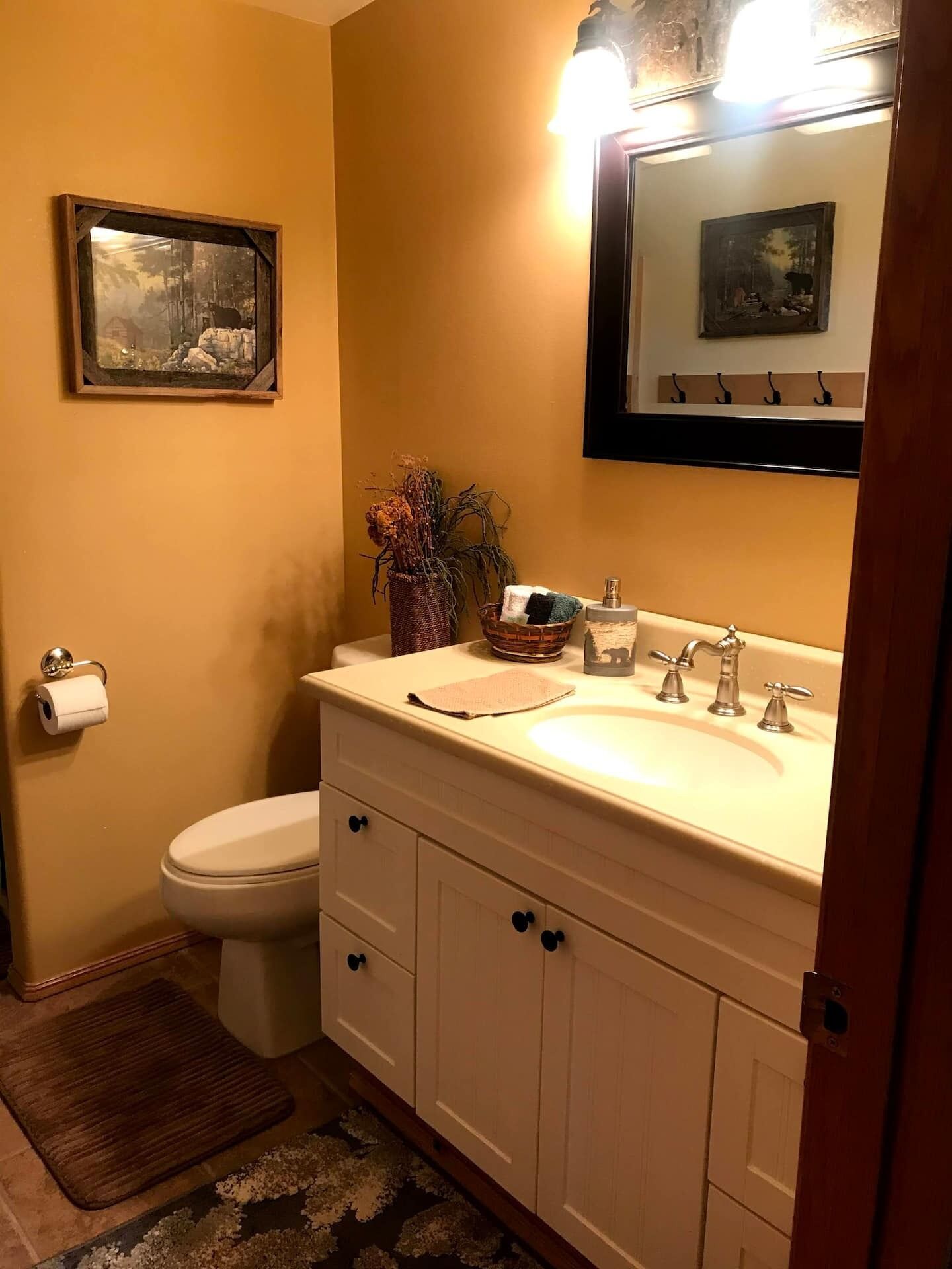 Baño con inodoro, lavabo y espejo. Hay un cuadro en la pared y un florero con flores secas sobre la encimera del lavabo.
