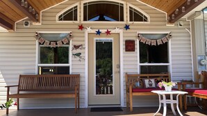 Eine Hütte mit Veranda, zwei hölzernen Bänken, einem kleinen Tisch mit Blumenvase und einer Tür, deren Schild „Cabin Sweet Cabin“ zeigt.