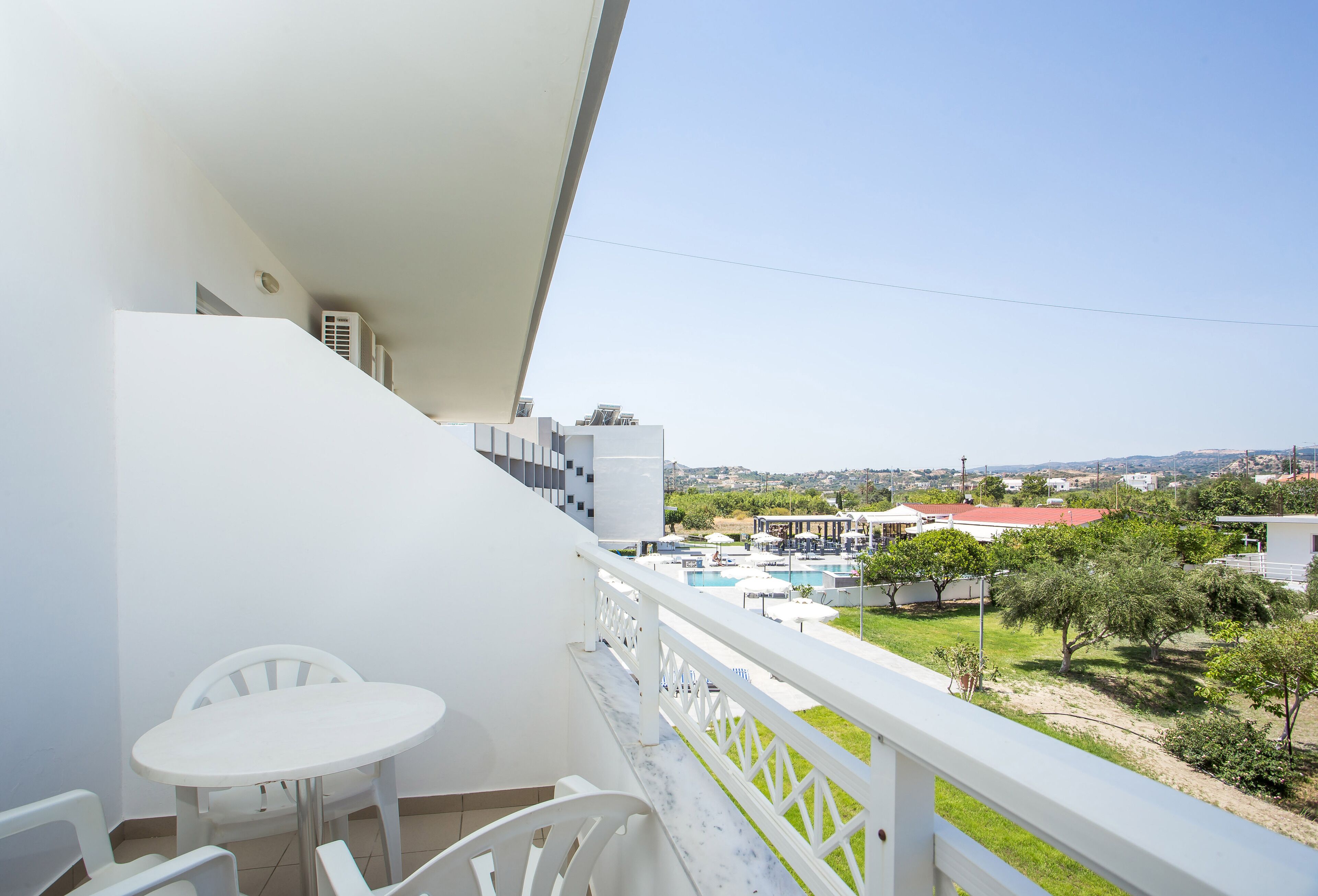 A balcony with a white table and chairs, overlooking a pool and greenery.