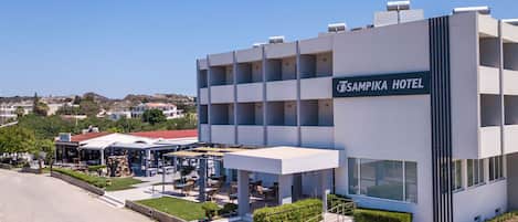 Un hotel moderno con un letrero que dice "TSAMPIKA HOTEL". Cuenta con una estructura de varios pisos, balcones y un área de estar al aire libre techada.